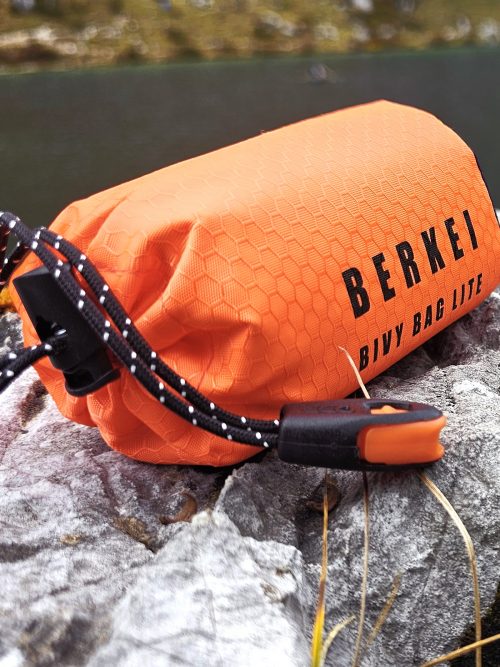 Leichter BERKEI Biwacksack Bivy Bag Lite in leuchtendem Orange auf Steinen am Wasser.