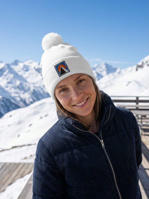 Junge Frau trägt das weiße BERKEI Bommel-Beanie Chalet in einer winterlichen Berglandschaft.